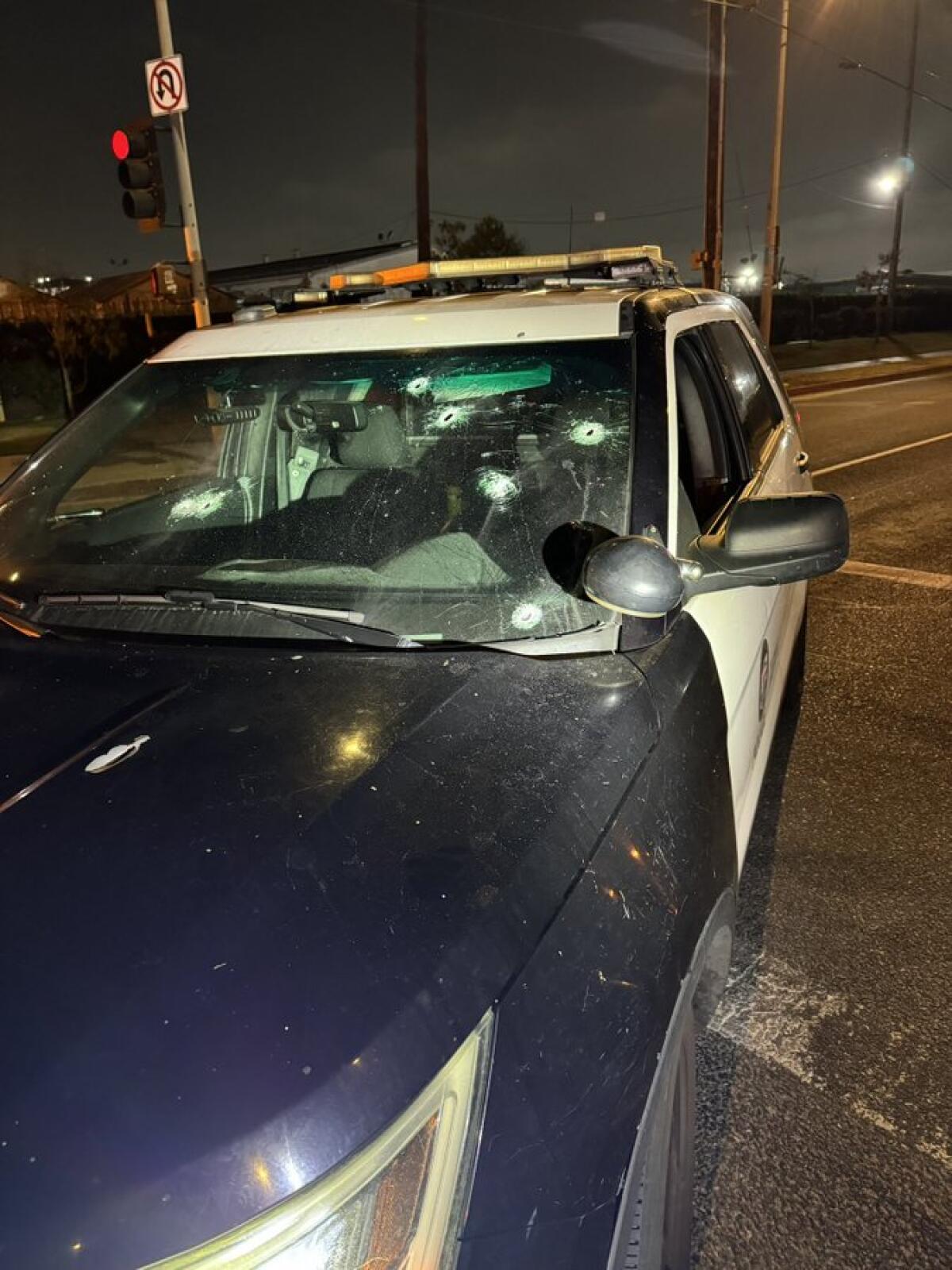 A stationary police cruiser on a street in the dark, its windshield riddled with bullet holes