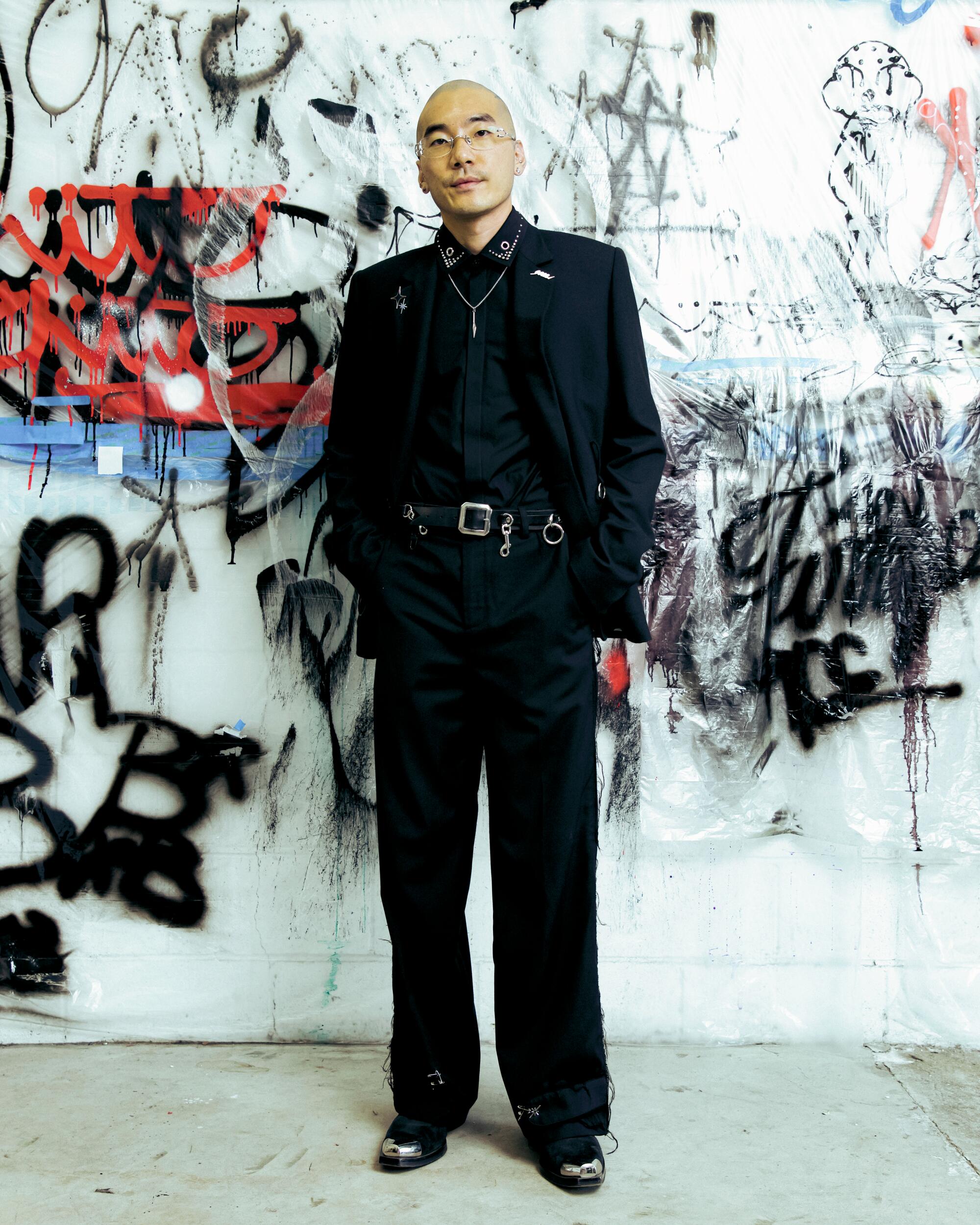 A man dressed in black stands against a white wall covered in colorful scribbles.