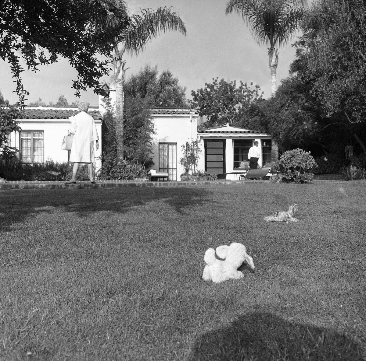 In the grassy yard of a home, a woman in dress with a purse on her arm stands in the background.