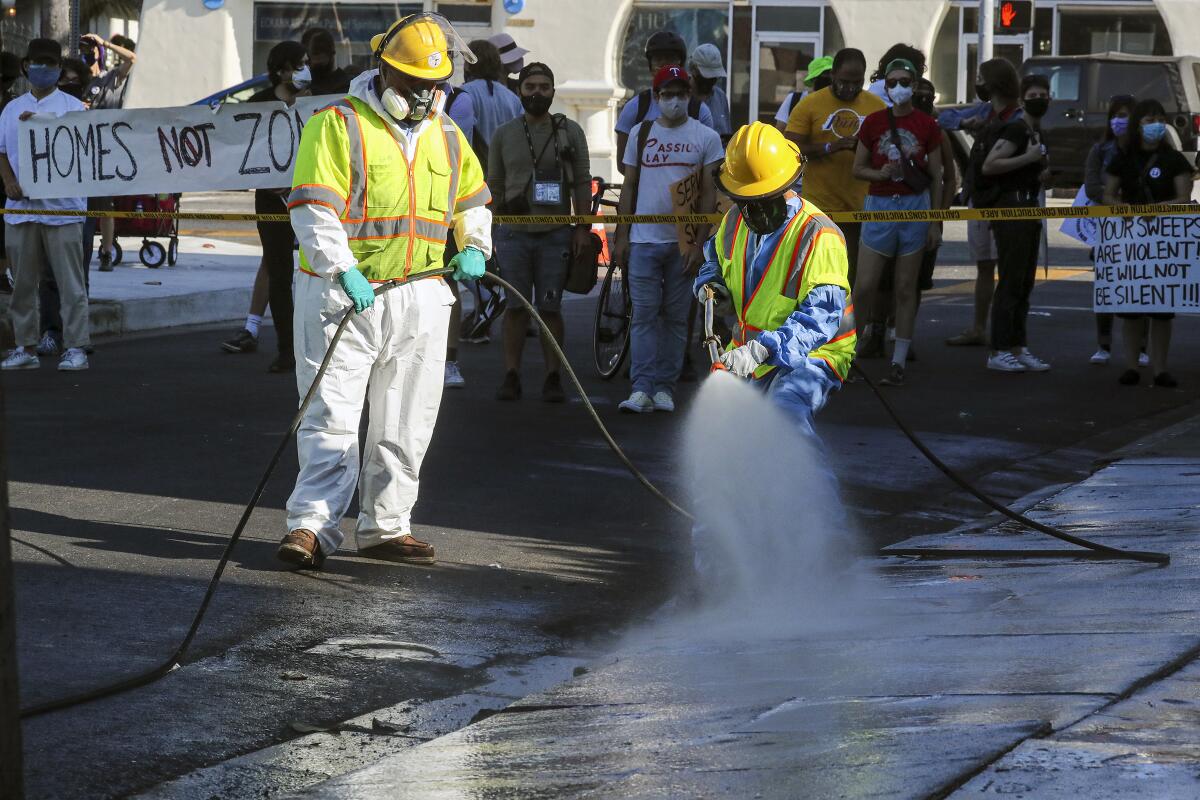 Workers in safety gear hose down a sidewalk.