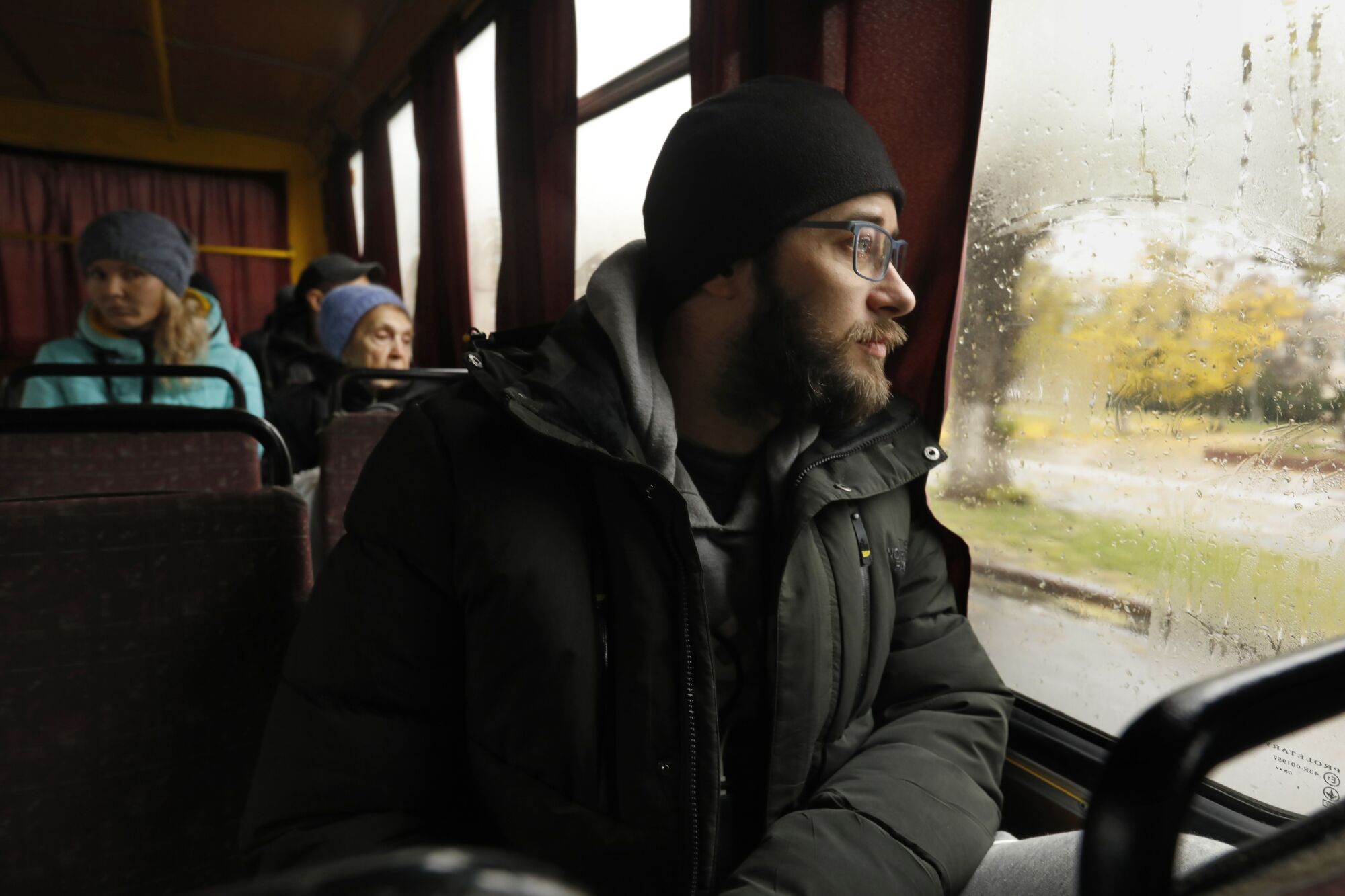 A man with a dark beard, wearing glasses and a dark cap and winter jacket, looks out the window of a bus