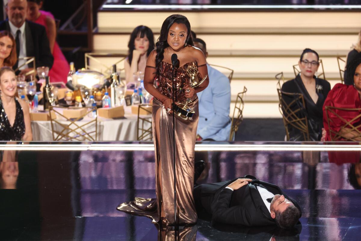 A woman in a gown holds an award and speaks into a microphone while a man in a tuxedo lies on the stage