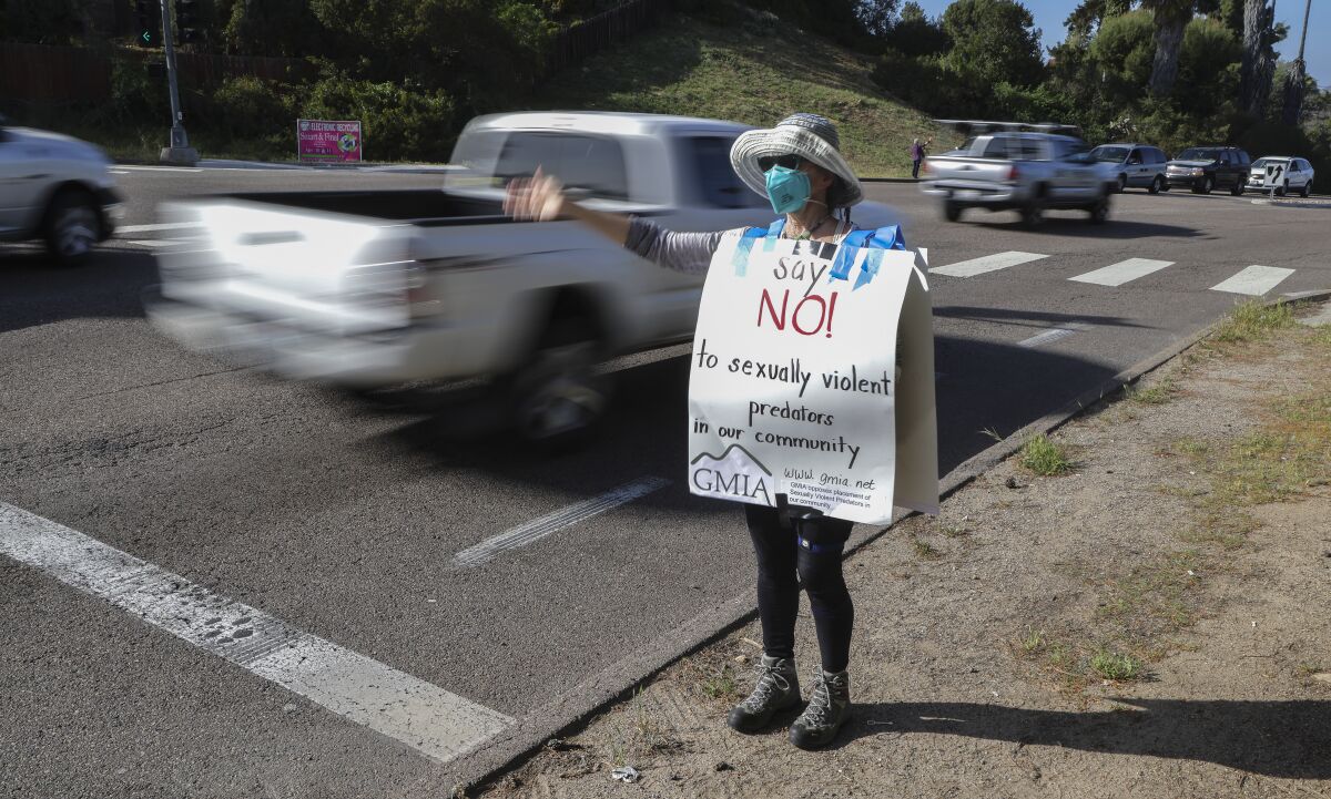 A woman waves to drivers on a busy road, wearing a sign that says "Say NO! to sexually violent predators in our community."