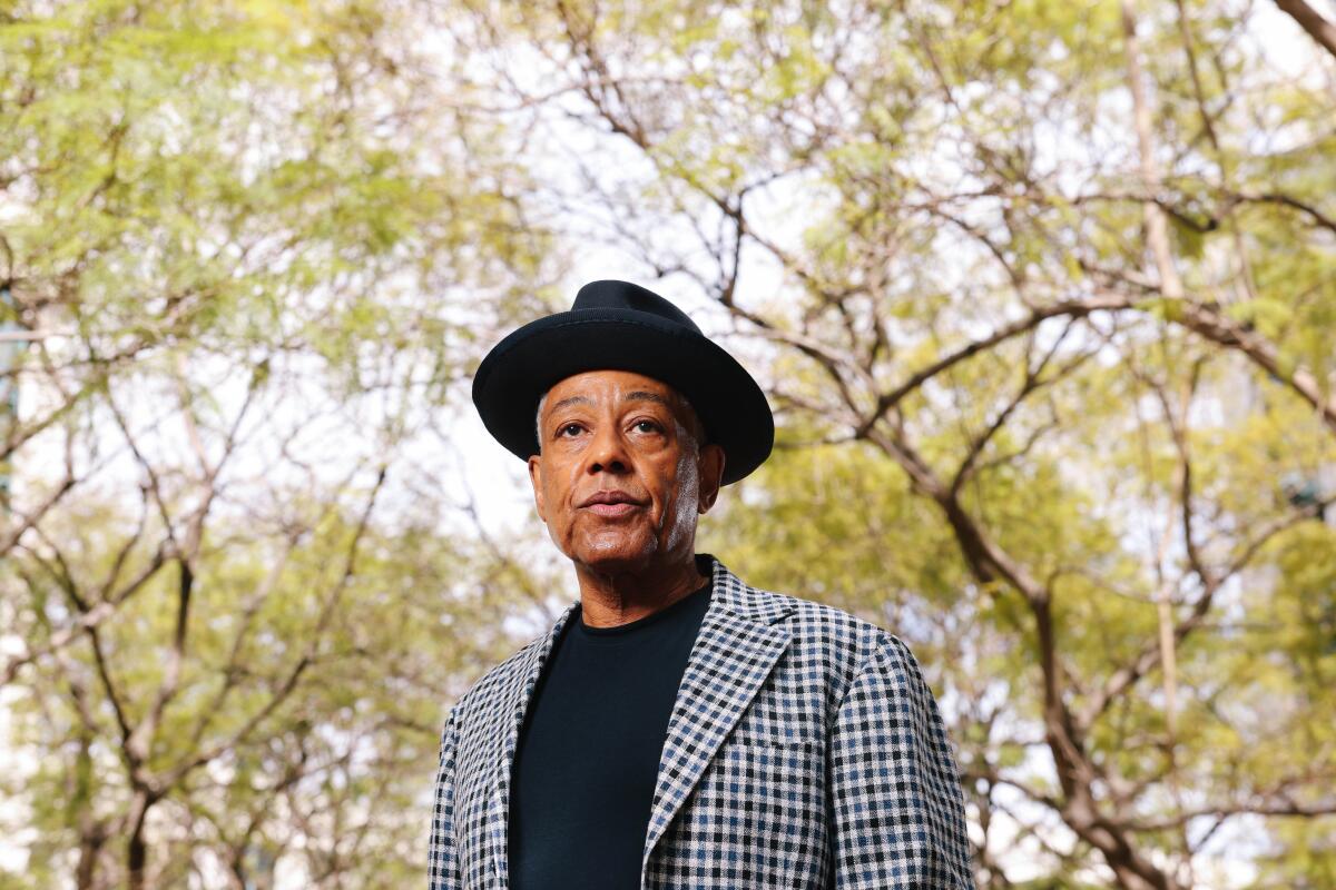 A man in a black hat and shirt with a checkered blazer standing with trees in the background.