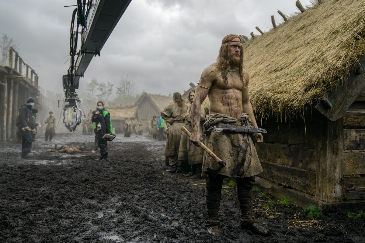 A Viking stands in the foreground of a village set while filming a movie.