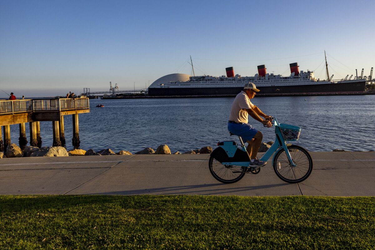 A bicyclist rides on Long Beach Shoreline Bike Path, with the Queen Mary visible across the water.