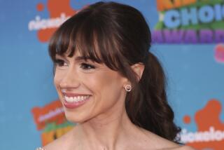 Colleen Ballinger is smiling while wearing a strapless pink dress with a thin shiny sash. 