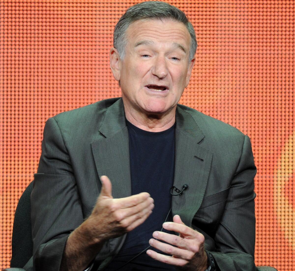 Robin Williams opens his mouth and gestures with his hands while sitting in front of an orange background.