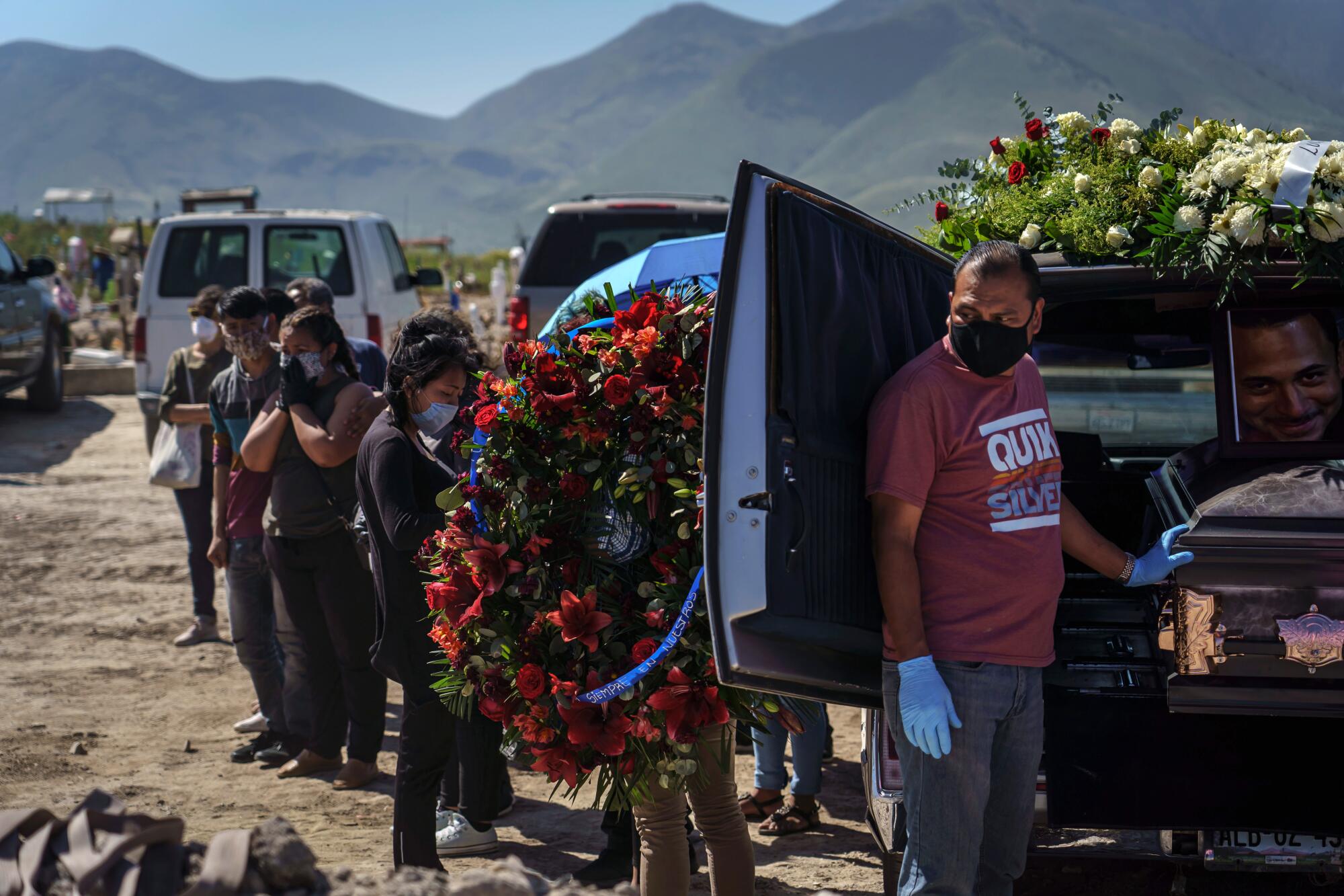 A funeral home worker prepares to unload a casket for cemetery workers to transfer into a grave as family members of the deceased stand nearby.
