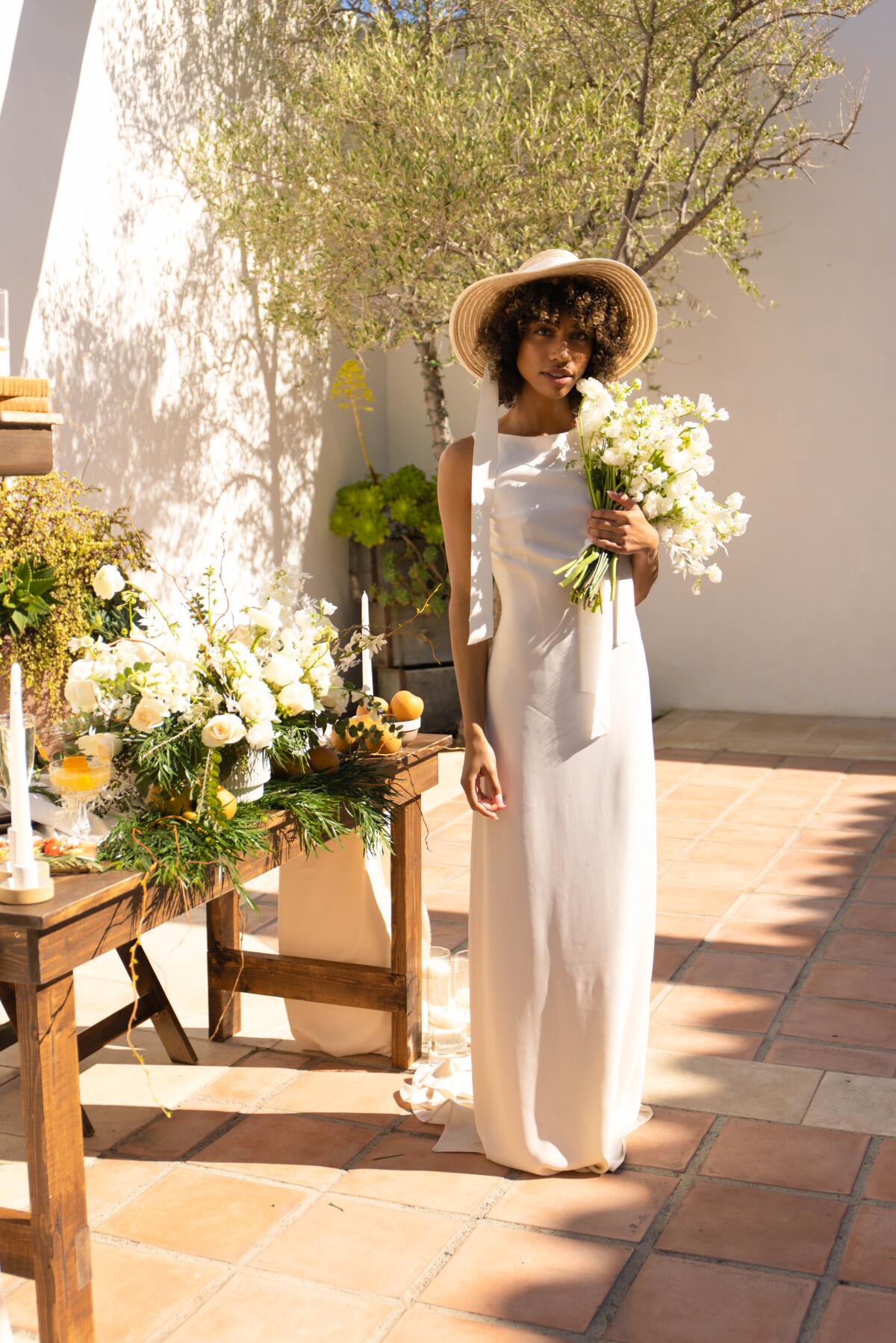 A model stands outside wearing a vintage Valentino cream dress and a hat and holding a large bouquet of flowers.