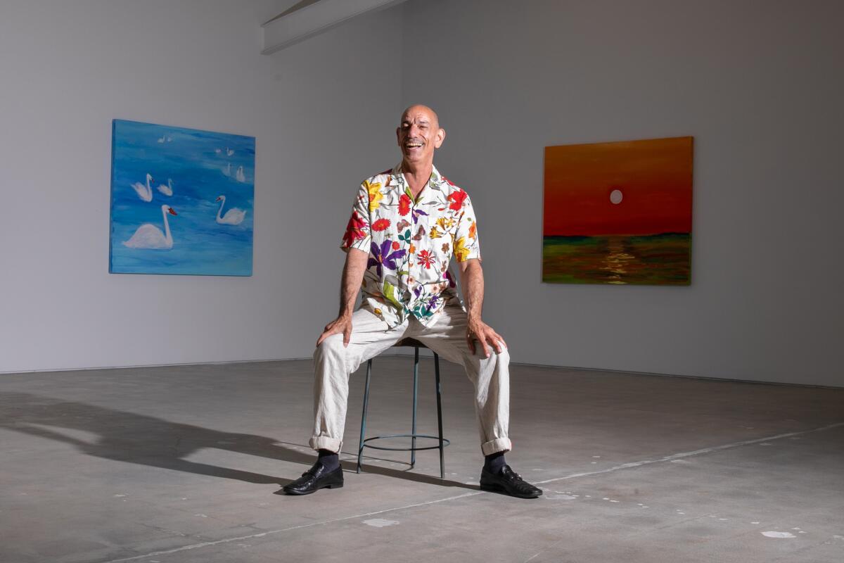 A man in a floral shirt poses in front of two paintings, one blue and the other orange.
