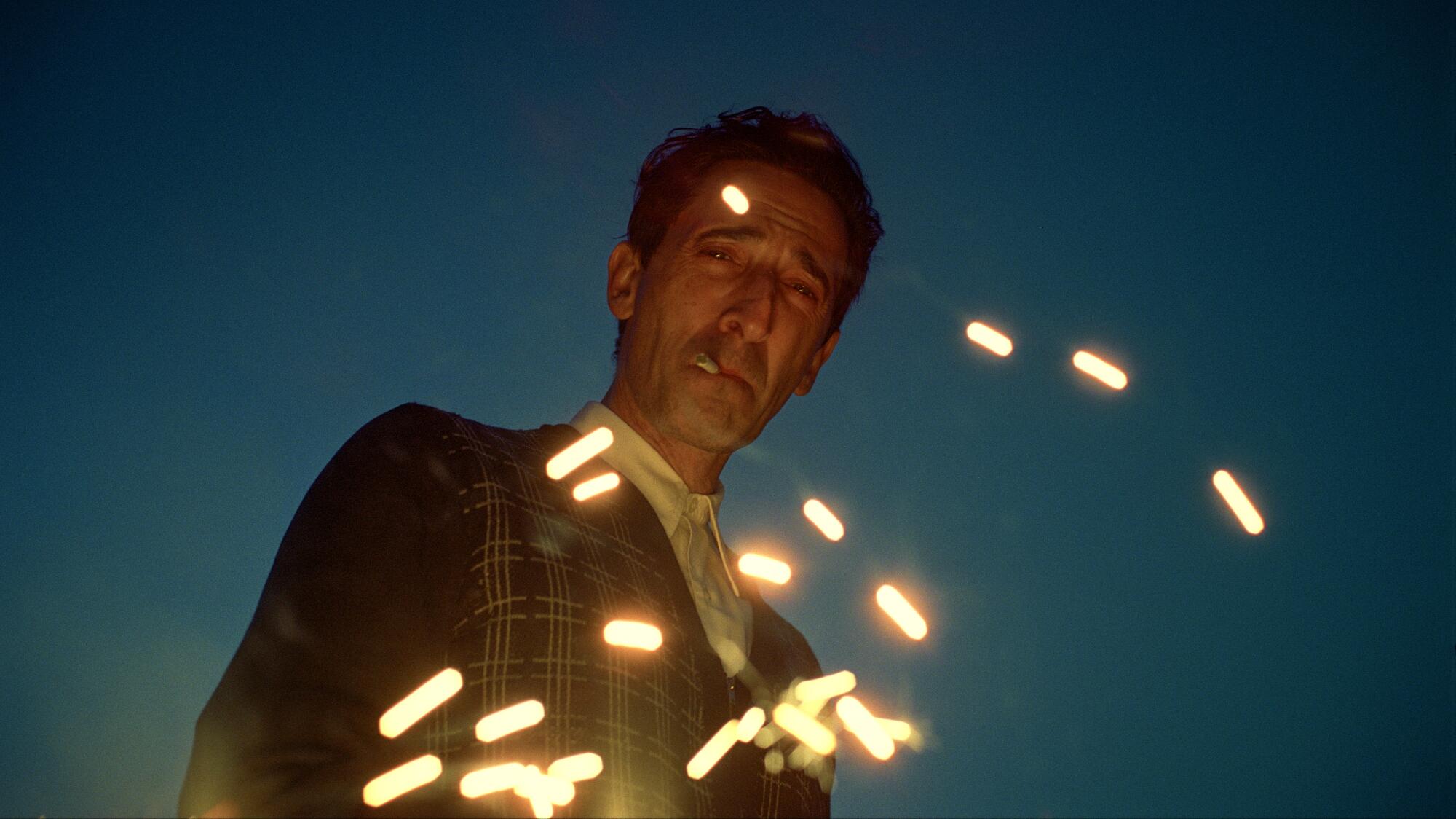 Adrien Brody smokes a cigarette and looks down as sparks rise up in "The Brutalist."