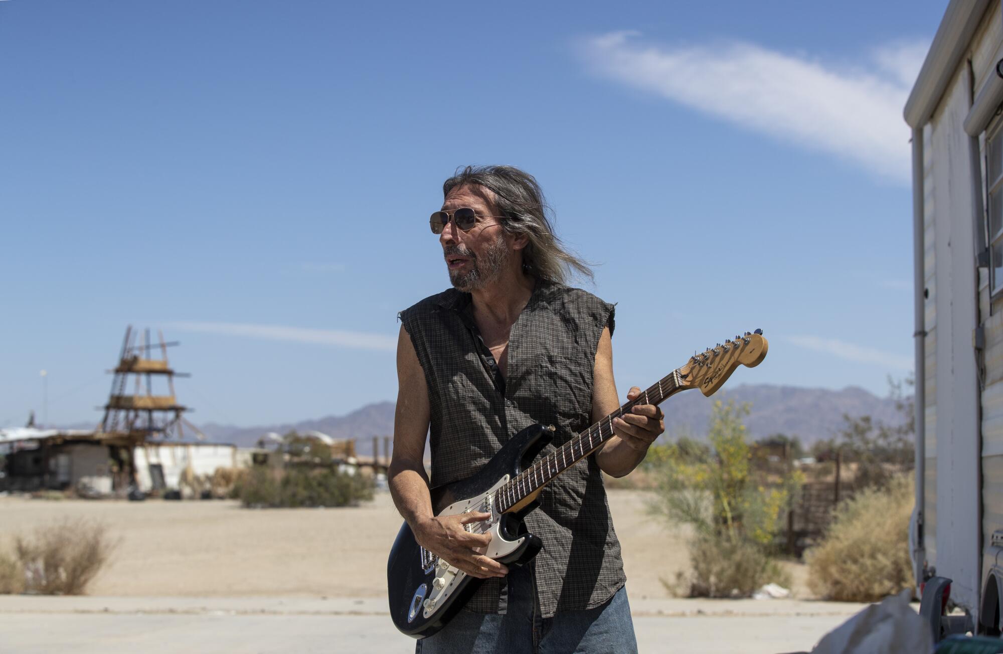 A man with long hair holds a Fender Stratocaster style electric guitar outside in the desert