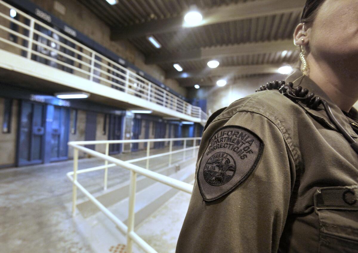 Closeup of a person in uniform standing inside a jail. Their shoulder patch reads "California Department of Corrections."