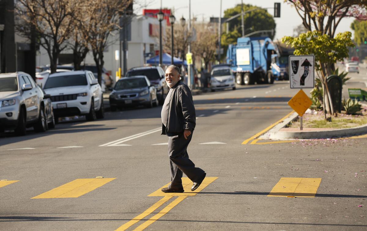 A man walks in a crosswalk marked in yellow near a street lined with parked cars  