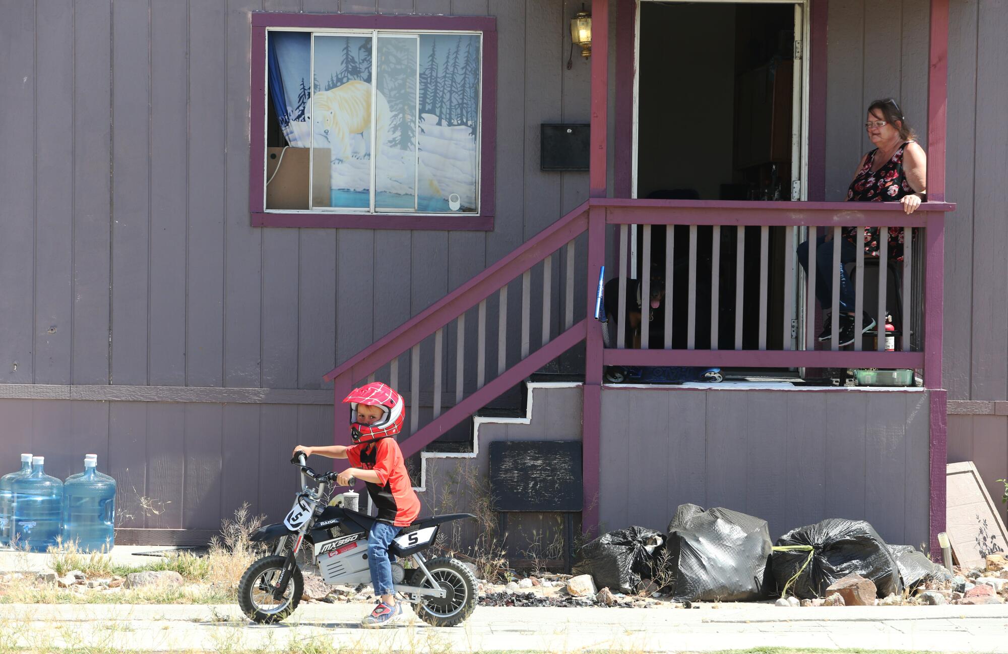 A woman leans on a porch railing as she watches a young boy on a bicycle.