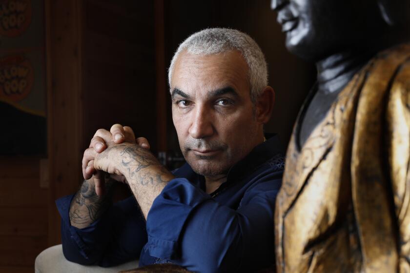 Alki David sitting in a chair and posing with his fingers laced together while wearing a blue collared shirt