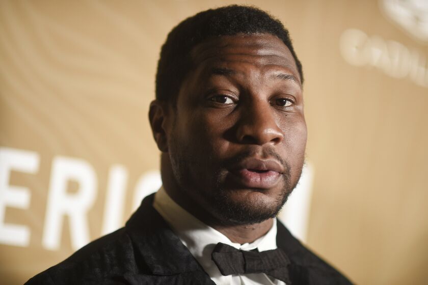 Jonathan Majors posing in a black suit and bowtie against a yellow background