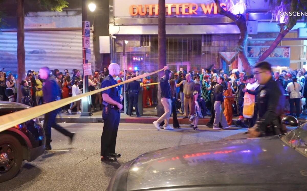 Police stand behind crime-scene tape near a crowded sidewalk.