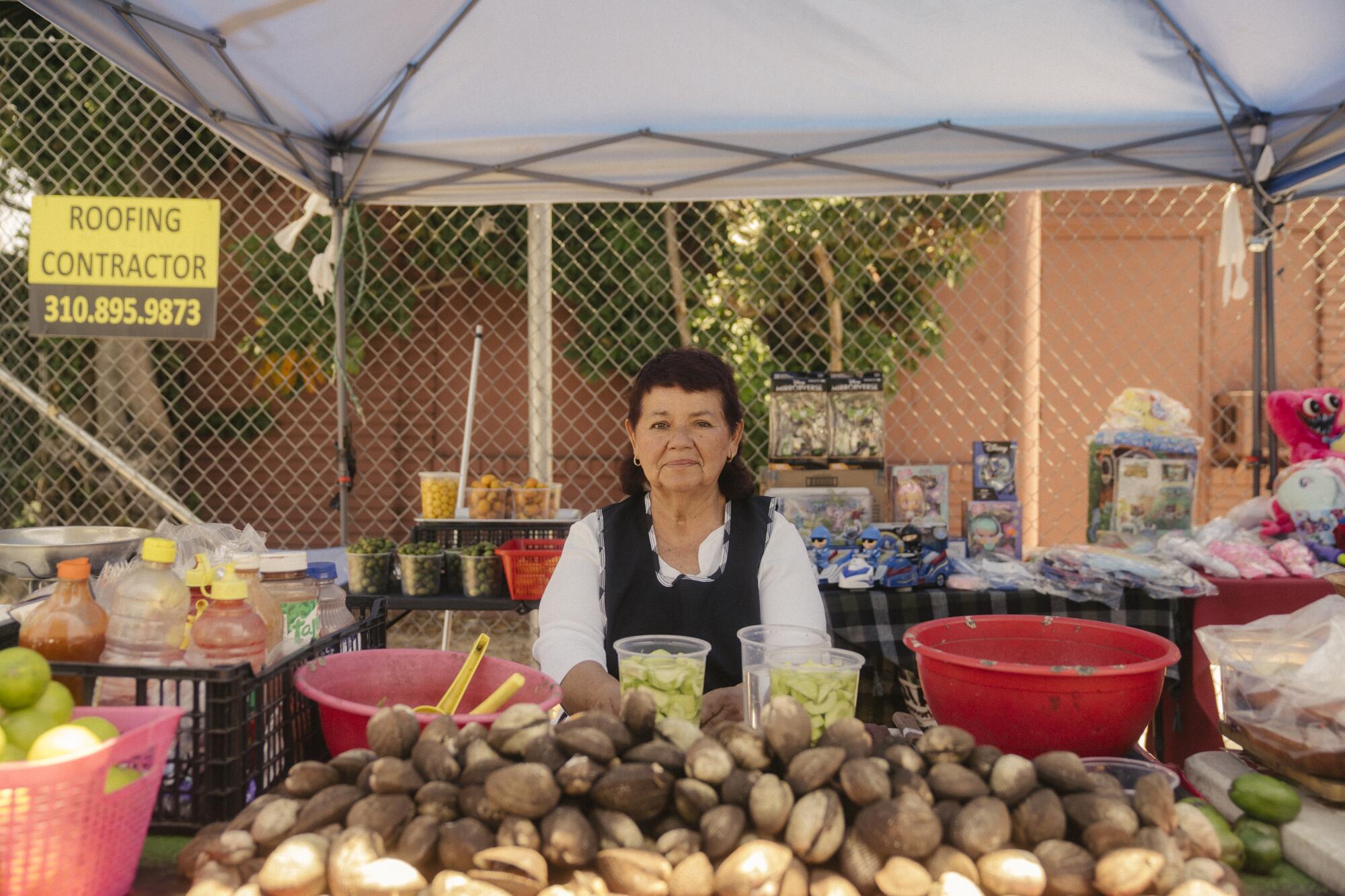 A woman stands behind an outdoor table selling food and drink, with a chain-link fence behind her