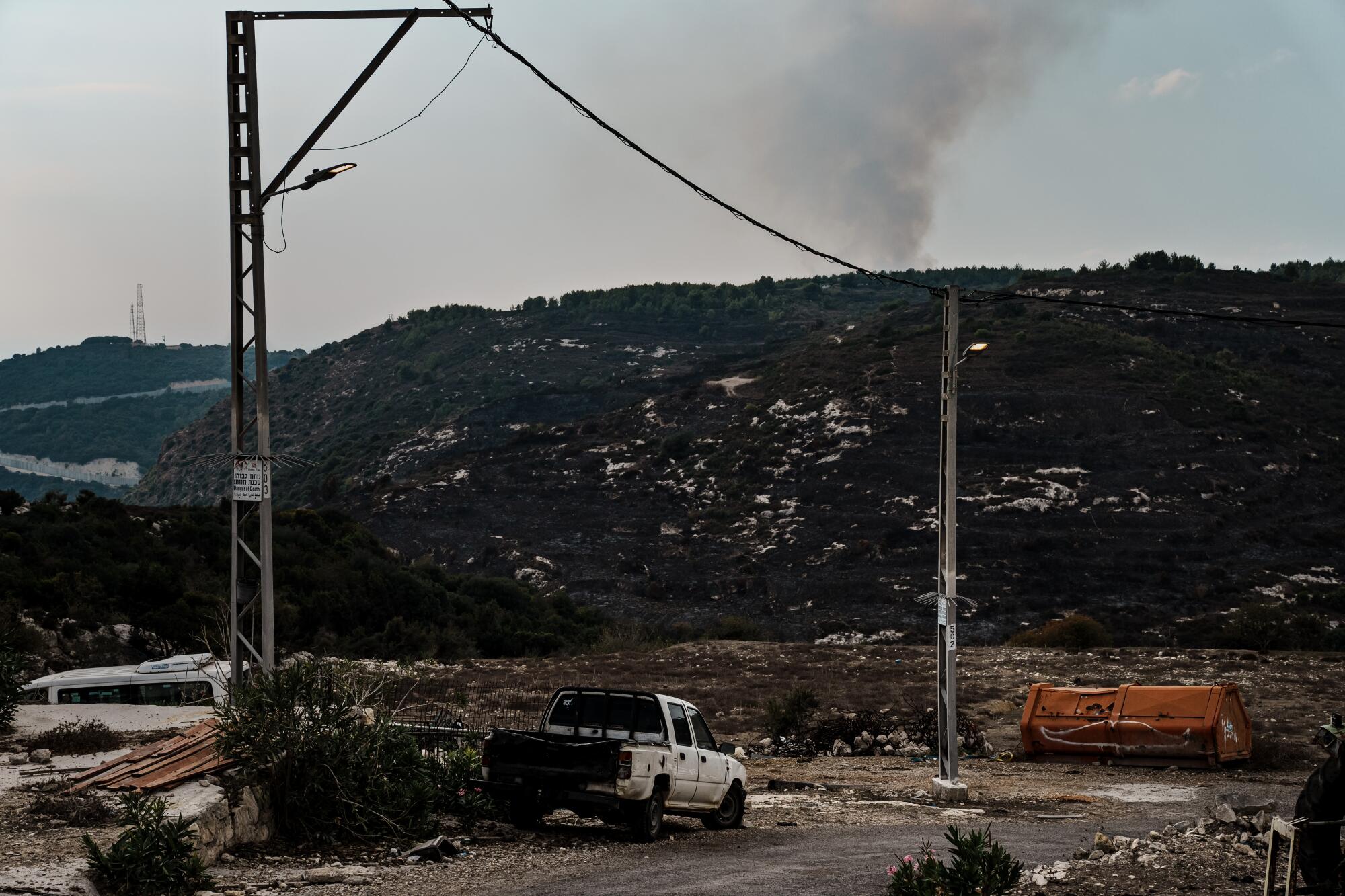 A plume of smoke rising from a hill serves as the backdrop to a brown landscape with white vehicles