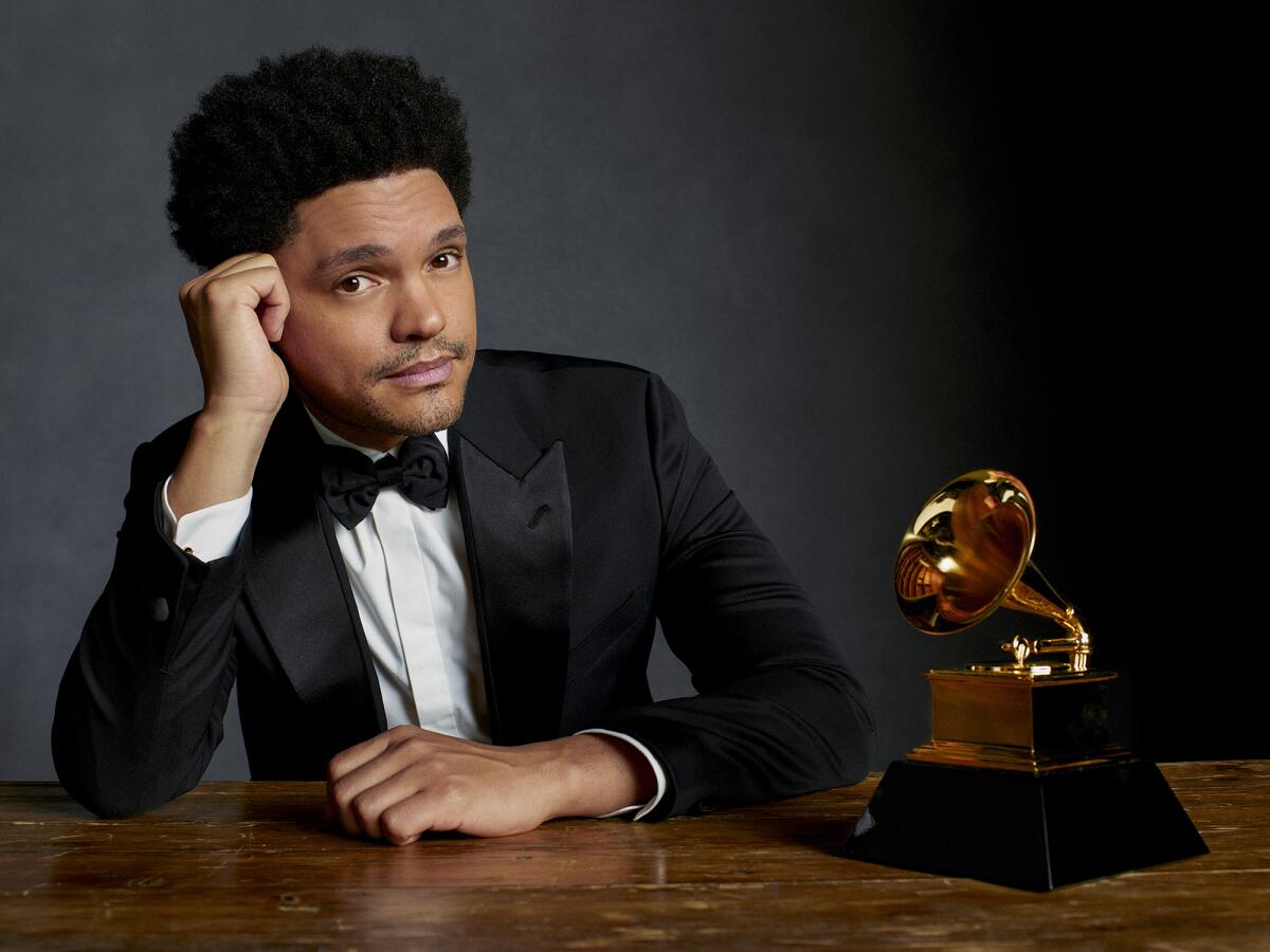 Trevor Noah posing in a tuxedo next to a golden Grammy trophy