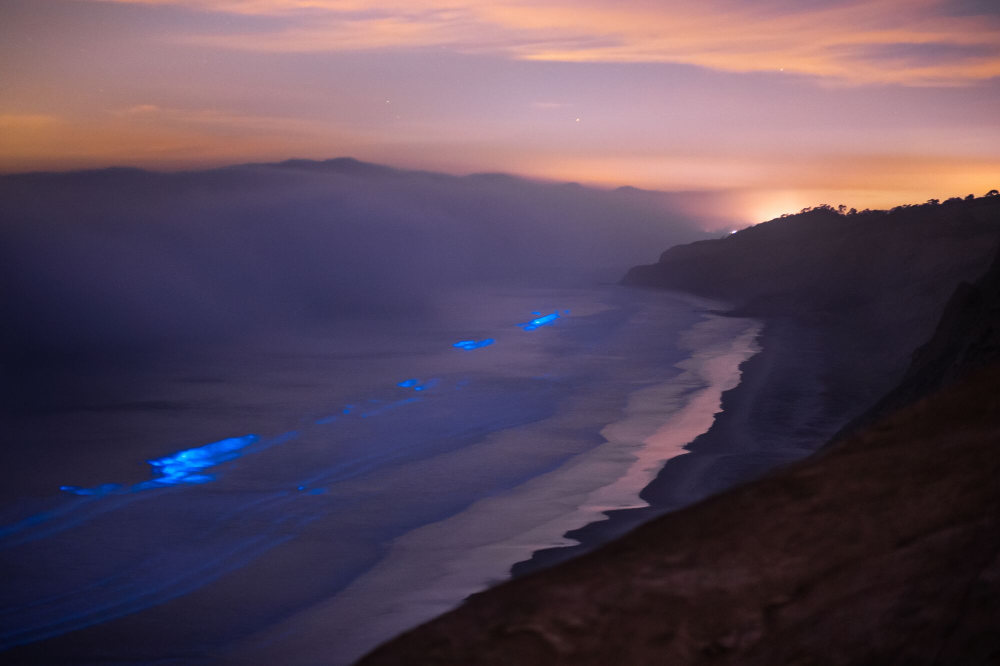 Bioluminescence off the coast while clouds sit above the water.