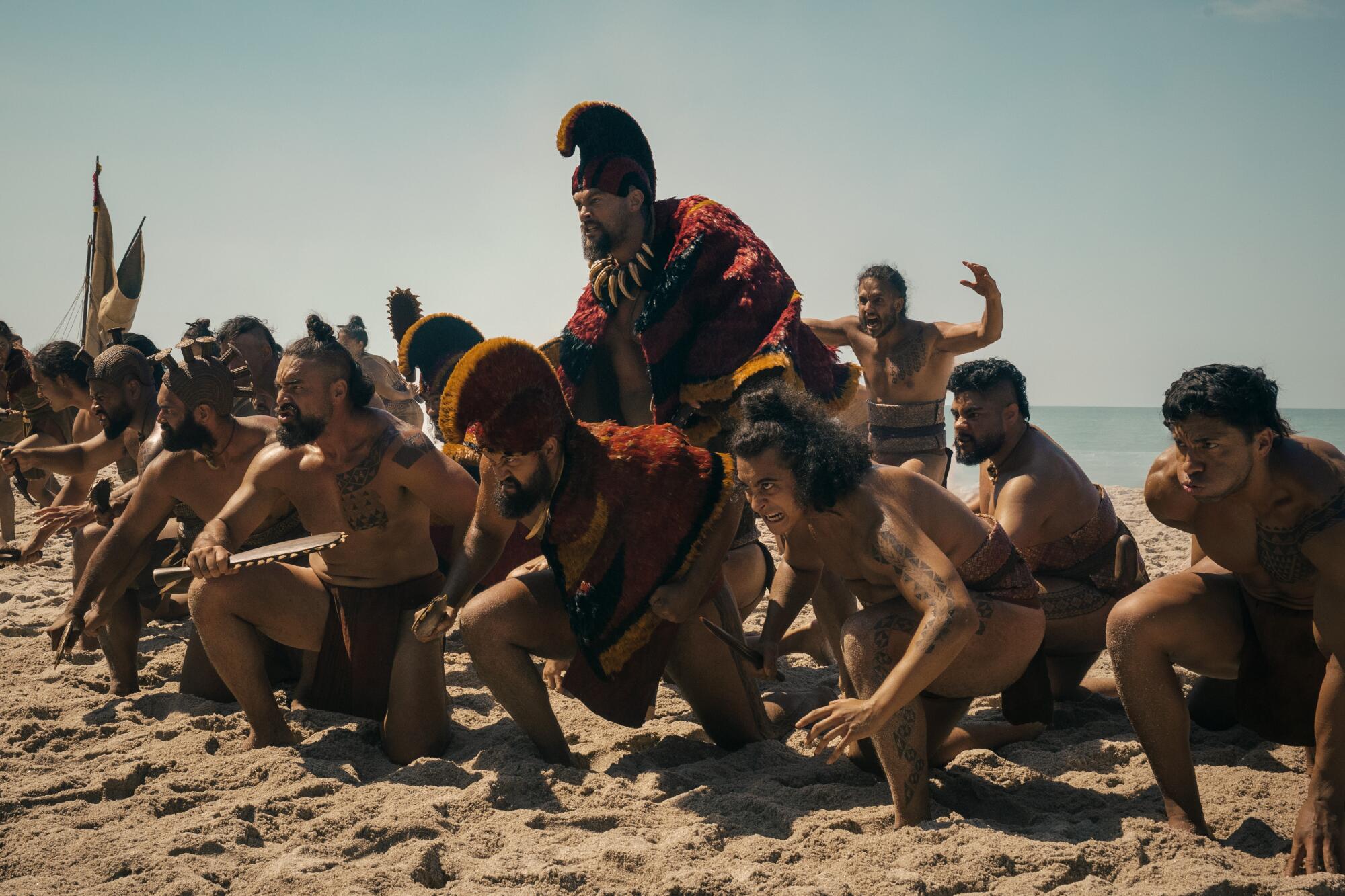 A group of men crouching in the sand on the beach with one man in a red headdress standing above them.