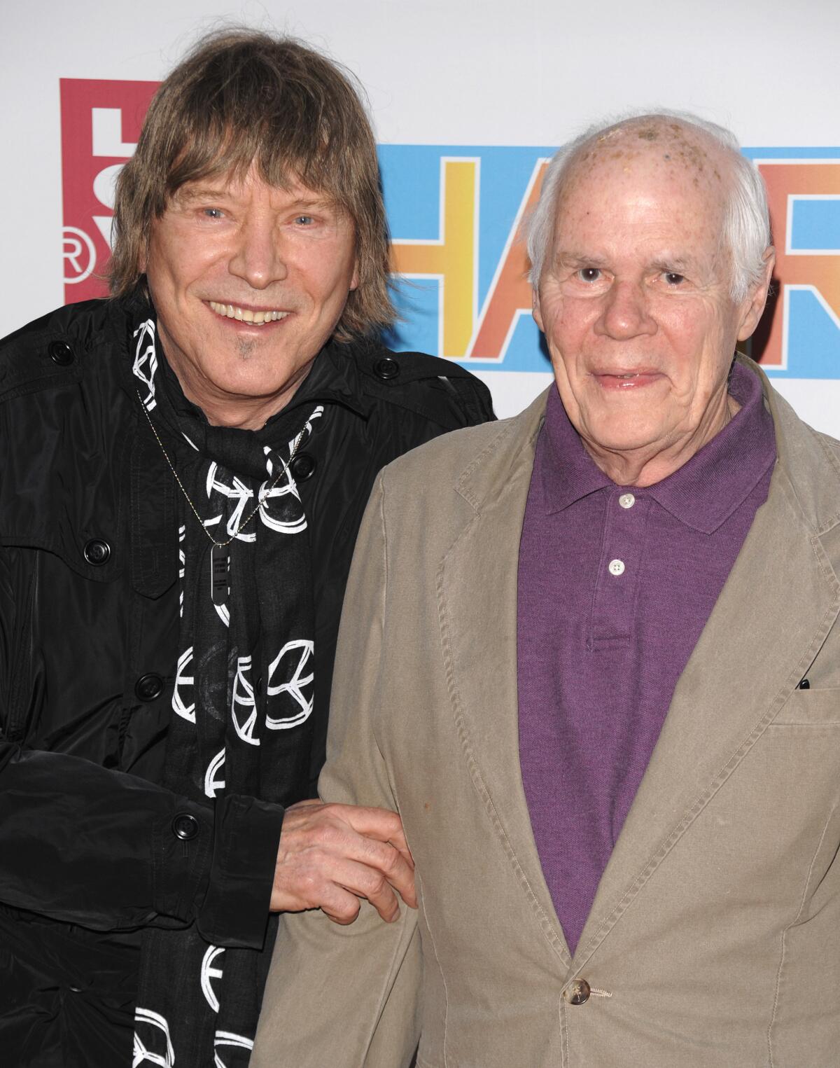 Two men smile for a photo. One wears a black shirt with peace symbols; the other is in a polo shirt and jacket.