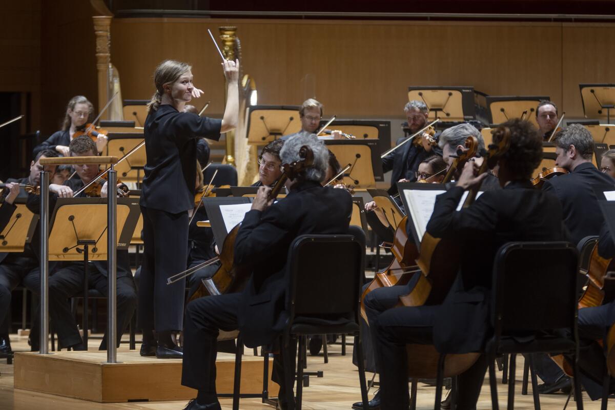 A woman wearing black stands on a conductor's podium and leads an orchestra.