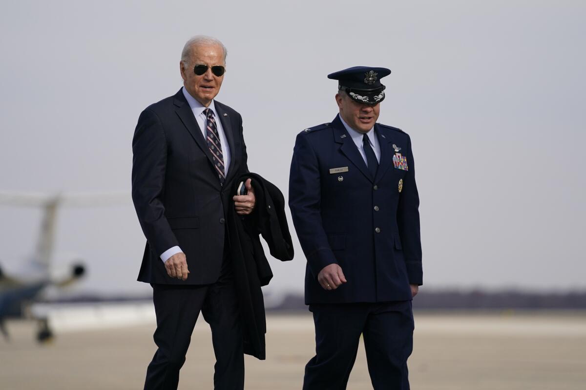 A man in dark glasses and dark suit and tie walks with another man in dark uniform and hat 