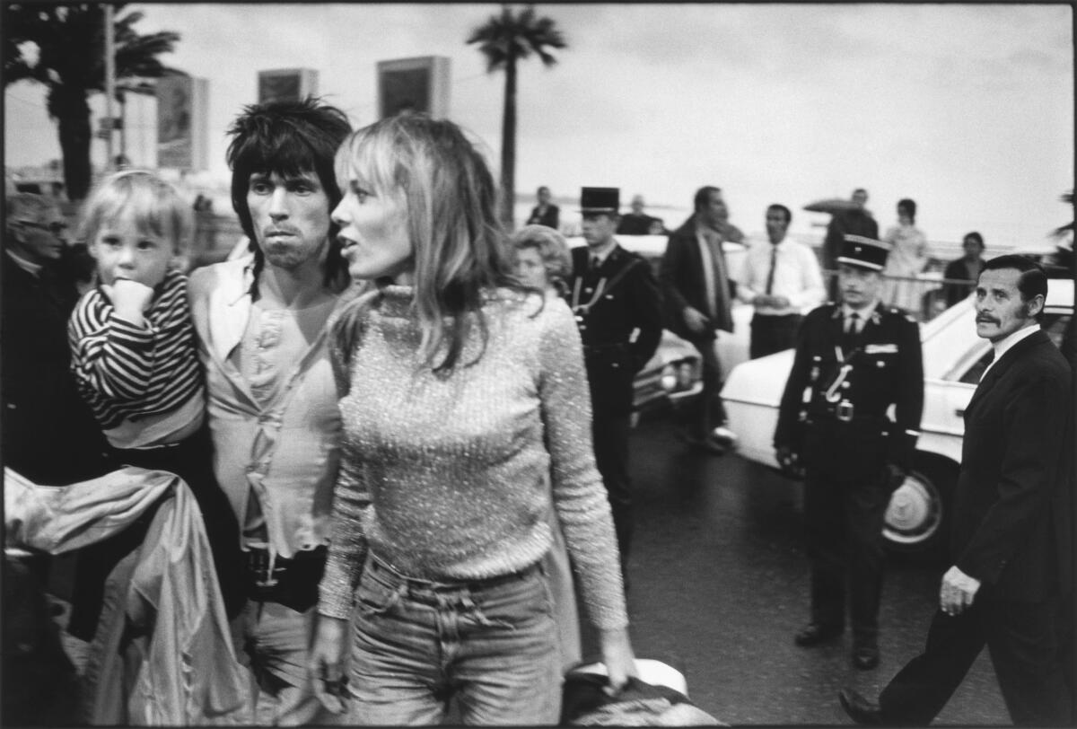 A man carrying a toddler and a woman speak to someone off camera as people mill around behind them