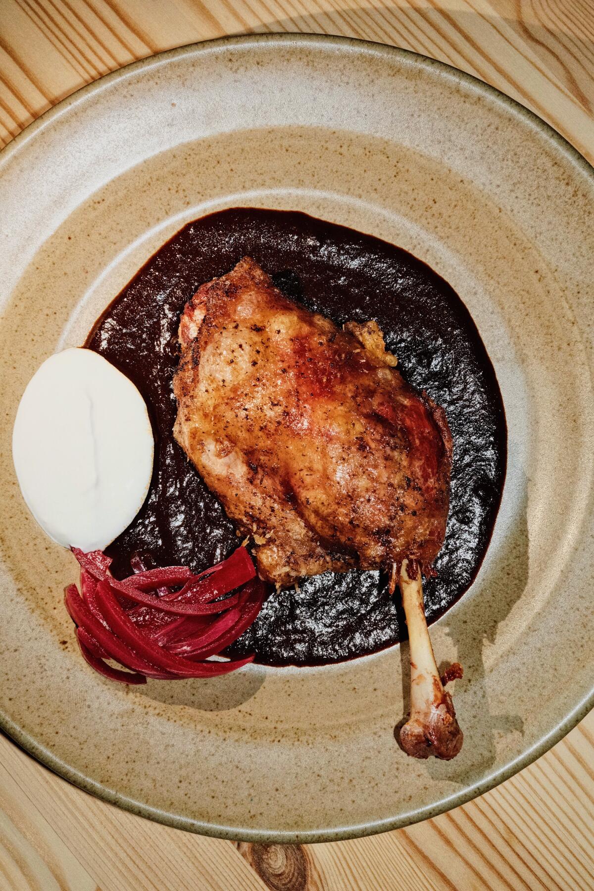 A bone-in duck leg with date mole, pickles and crema on a ceramic plate on wood table at A Tí in Echo Park