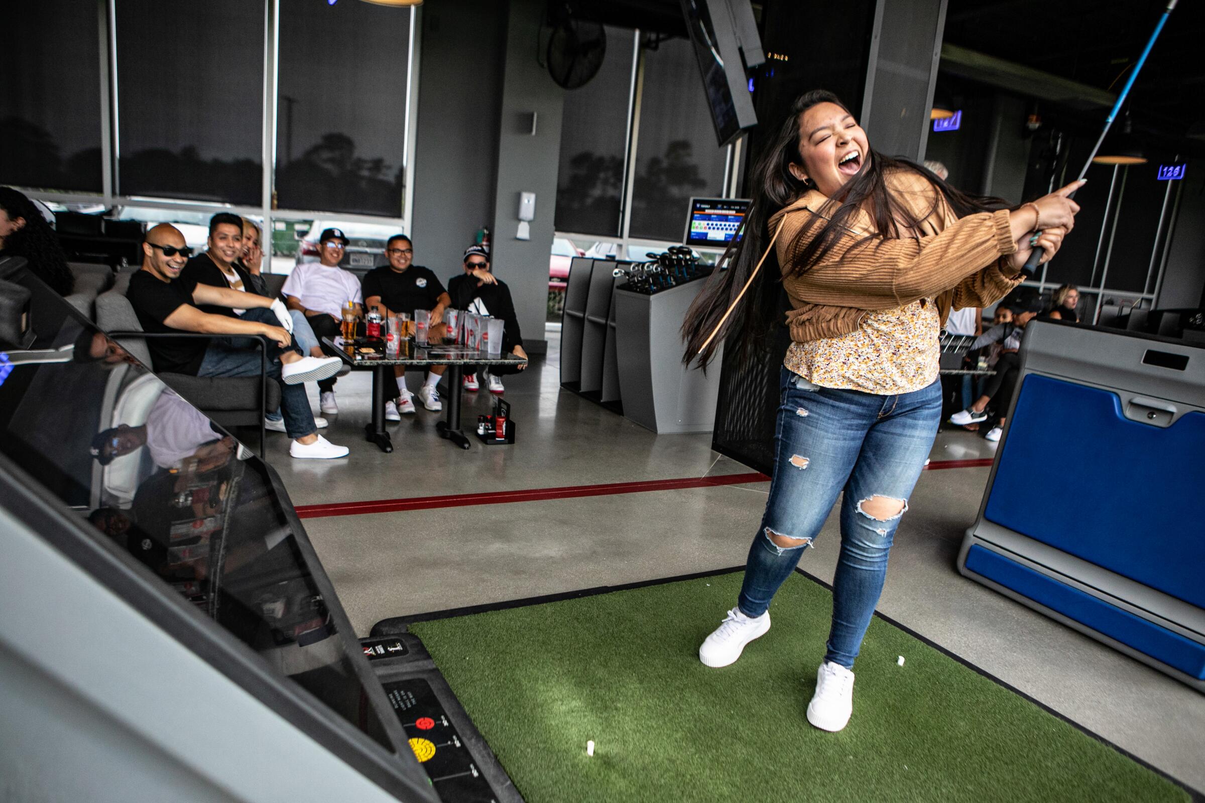 A woman swings a golf club indoors as friends look on at Topgolf. 