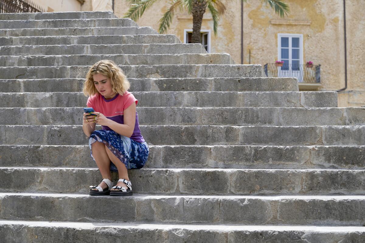 A blond woman looking at her phone sits on steps outside.