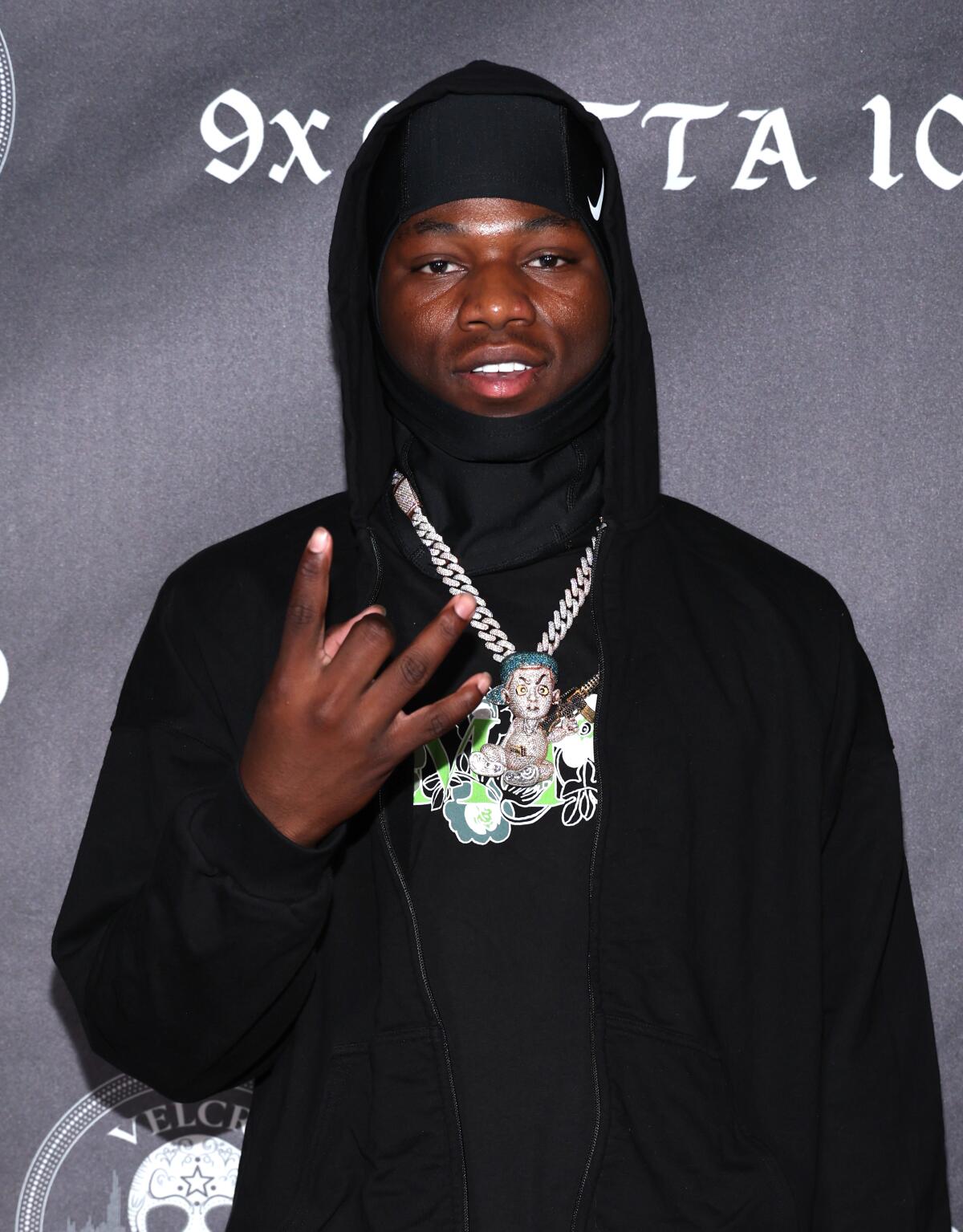 Trench Baby flashes a hand sign while wearing a black hoodie and a silver chain