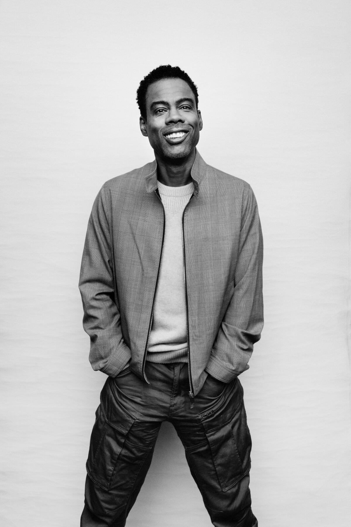 A black and white photo of Chris Rock, wearing a jacket, T-shirt and jeans and standing with his hands in his pockets.