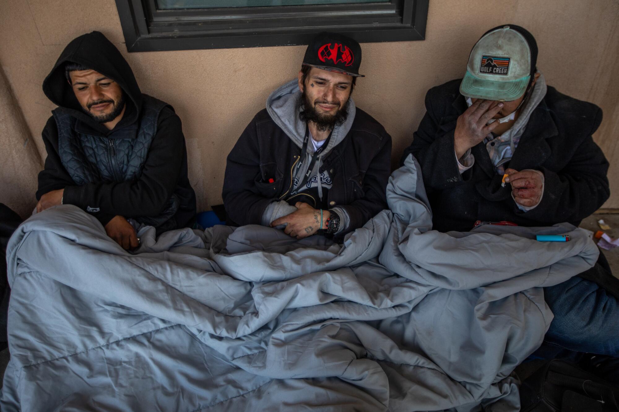 Three men sit outdoors sharing a blanket