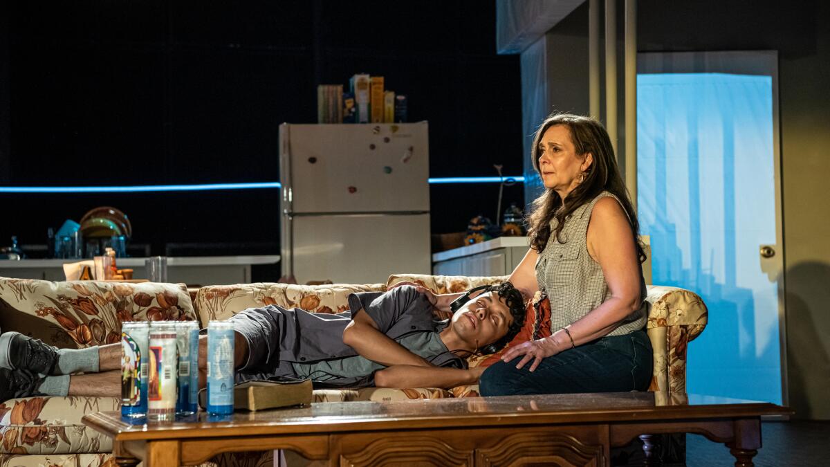 A woman sits on a sofa with a young man lying on the sofa in a stage-set apartment.