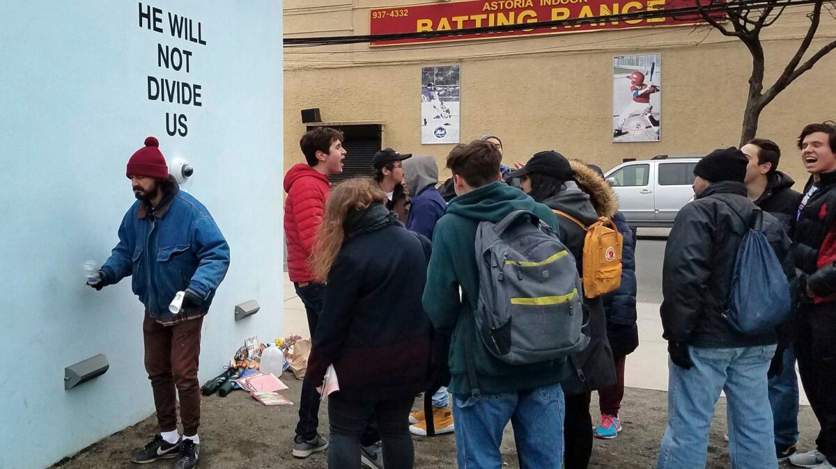 Actor Shia LeBeouf, left, stands in front of a live-steam camera with the words "He Will Not Divide Us" posted on a wall outside of the the Museum of the Moving Image in New York.