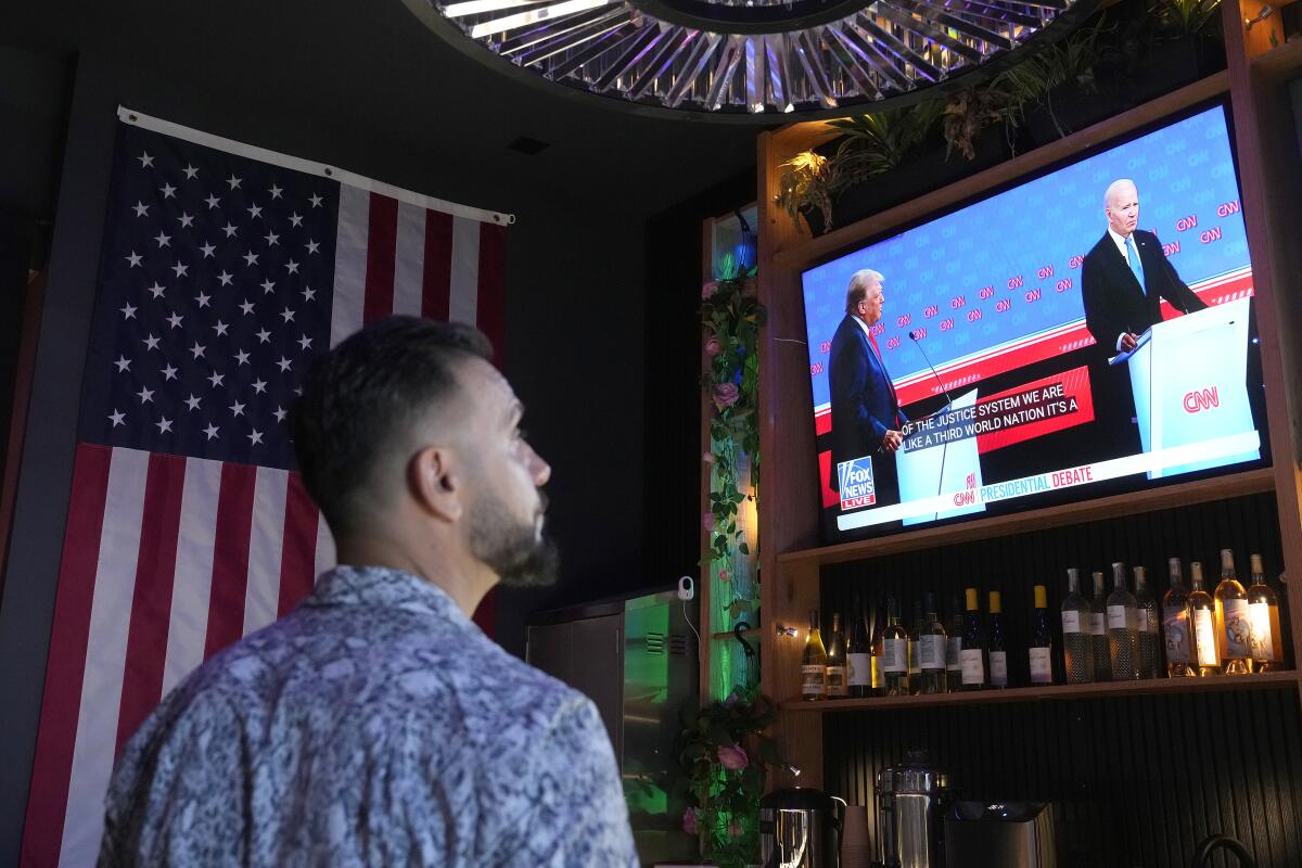 A bearded man in a collared shirt watches the CNN presidential debate on a TV above an alcohol shelf next to an American flag