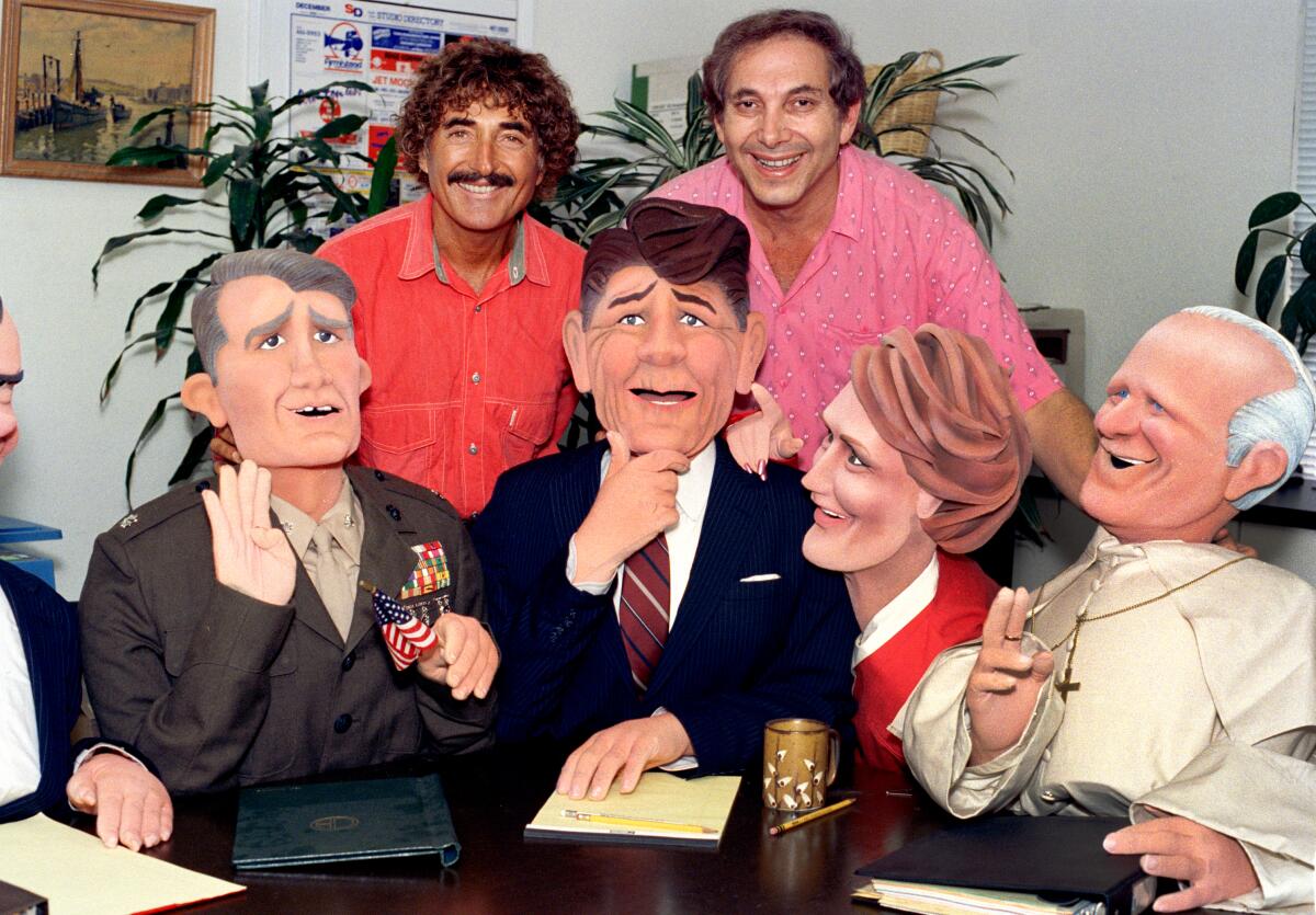 Sid Krofft and Marty Krofft stand behind four life-size puppets in suits seated at a table.