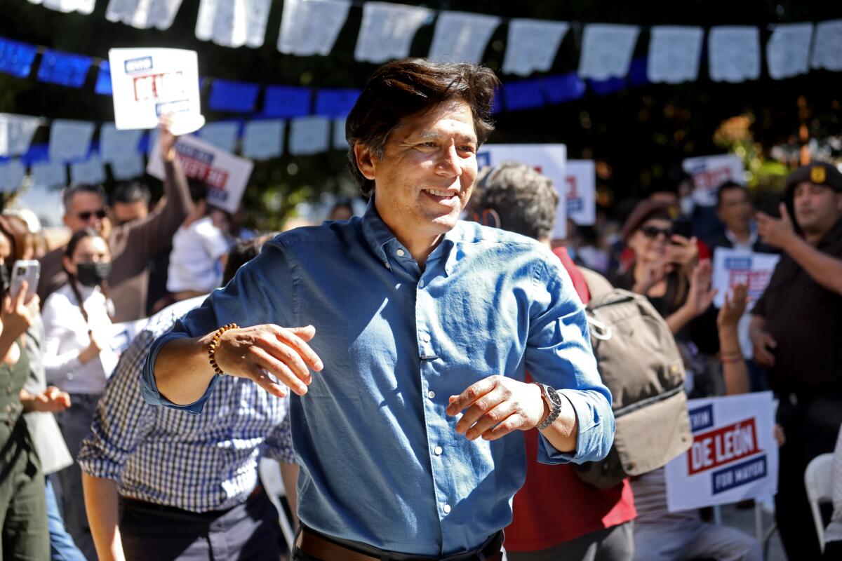 A smiling man in a blue button-down shirt walks among a crowd of people, some in masks.