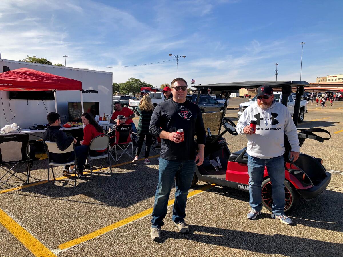 Two men hold cans of beer as people tailgate in a parking lot