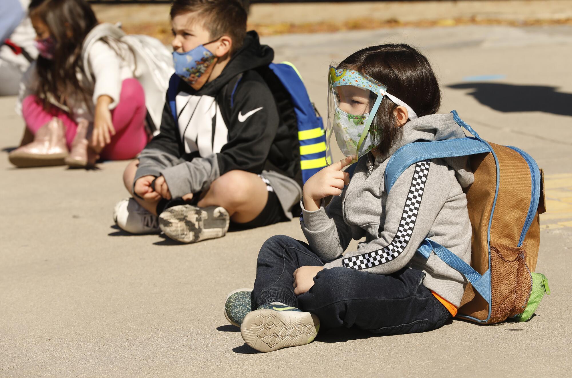 Students wearing backpacks, including a young girl in face mask and shield, sit crosslegged outside school.