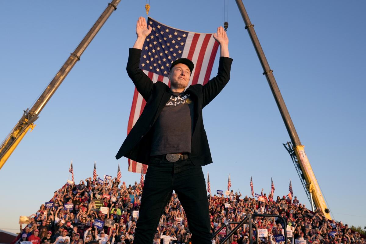 A man in dark clothing holds up his hands as he stands in front of an American flag, with a crowd in the background