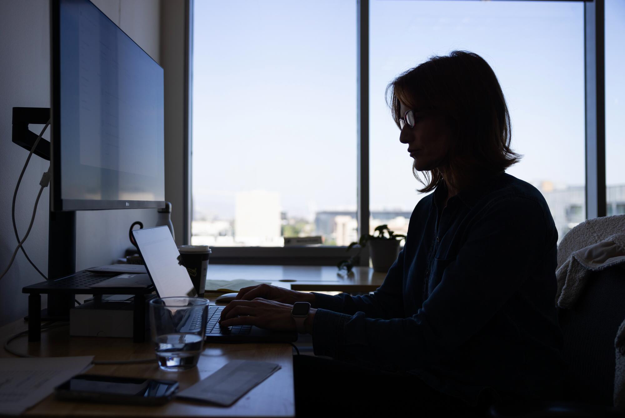 A woman at a desk in an office is silhouetted by light from large windows.