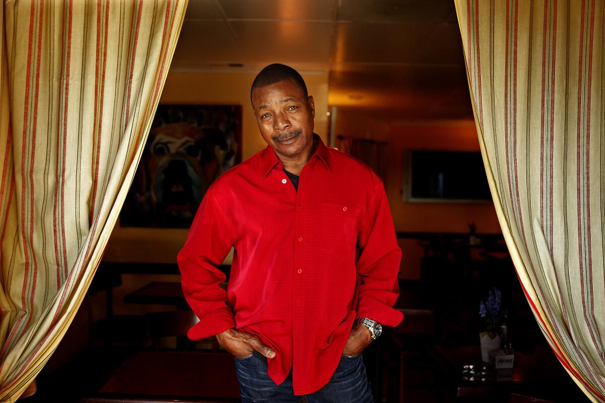 Oct. 2014 photo of Carl Weathers, wearing a deep red button-down shirt and standing between two curtains