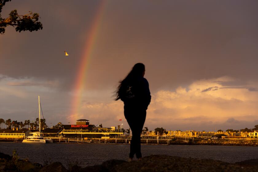 Long Beach, CA - March 15: A rainbow appears over Ballast Point Brewing Long Beach amid light rain as Ashley Contreras, of Long Beach, takes in the scene after a long day teaching school at the Alamitos Bay Inlet jetty in Long Beach Friday, March 15, 2024. (Allen J. Schaben / Los Angeles Times)