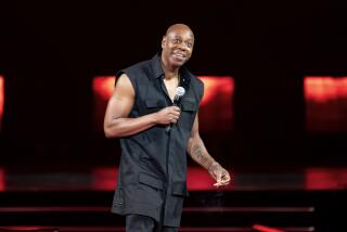 Comedian Dave Chappelle wears a sleeveless jacket and holds a microphone onstage